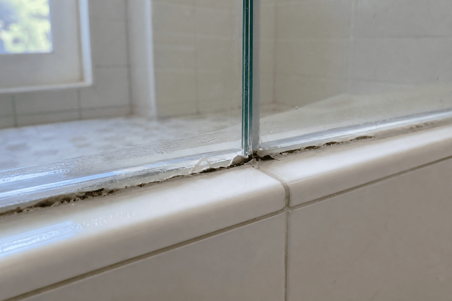 Cracked, peeling silicone with dark mold along the base of a frameless shower glass panel.